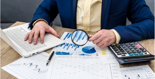 An analyst examining financial charts with a magnifying glass beside a laptop and calculator